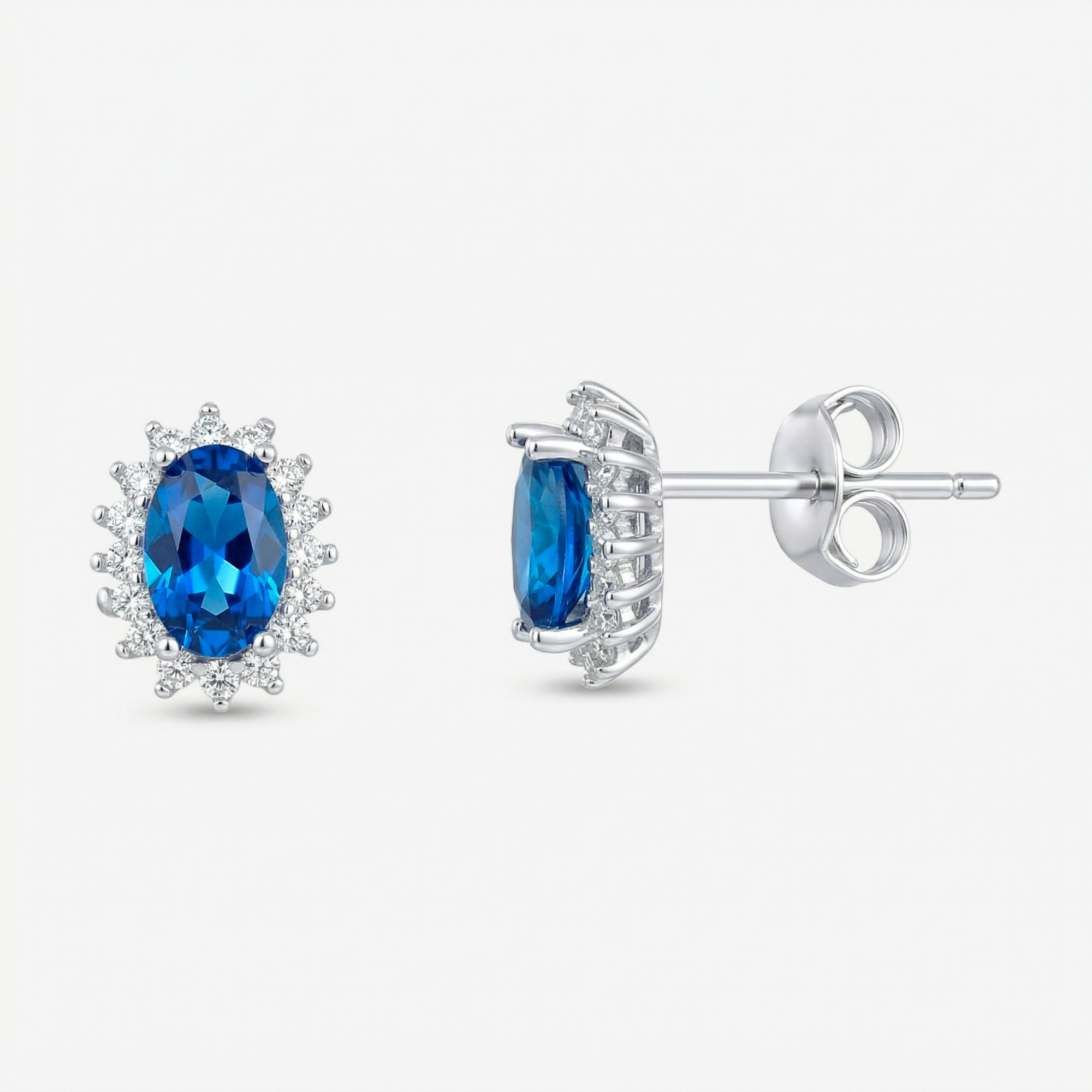 Par de brincos Avel em prata 925 com zircônia oval azul intenso em quatro garras e halo de zircônias brancas, vista frontal e lateral com fecho tarracha
