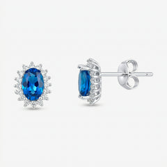 Par de brincos Avel em prata 925 com zircônia oval azul intenso em quatro garras e halo de zircônias brancas, vista frontal e lateral com fecho tarracha
